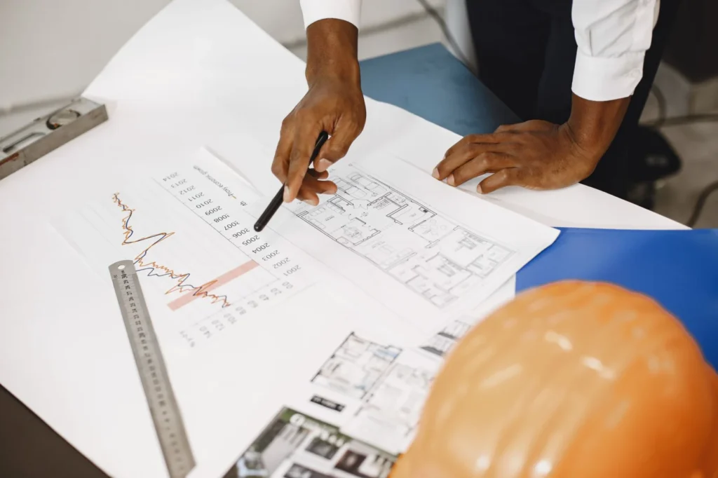 Hands of an architect analyzing blueprints and financial graphs at a desk.