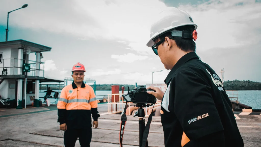 Two construction workers in safety gear filming at a waterfront location.